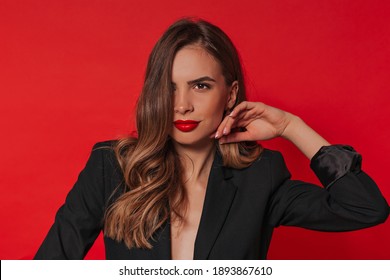 Smiling fashionable european woman with curly light-brown hair, wearing black jacket and red lipstick posing over isolated red background.