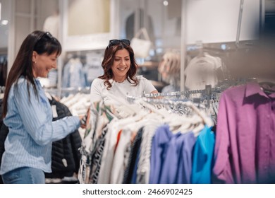 Smiling fashionable friends standing at store near rack with clothes and choosing dresses. Young trendy female customers at boutique buying and purchasing dresses and clothes on retail. Shopaholics. - Powered by Shutterstock