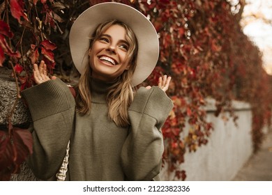 Smiling Fashionable Young Tanned Woman In Beige Hat Among Autumn Foliage Outdoors In City Park Autumn. Teenager Smiles Toothy Waving His Arms. Positive Emotion Concept