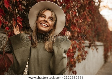 Smiling fashionable young tanned woman in beige hat among autumn foliage outdoors in city park autumn. Teenager smiles toothy waving his arms. Positive emotion concept