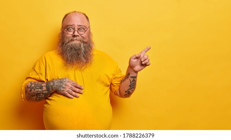 Smiling fat bearded man in eyewear with bald patch and tattooed arms, points on empty space aside, has big stomach after drinking beer, promots something on yellow background, recommends product