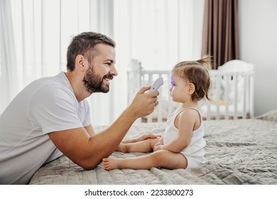 A smiling father is checking on temperature on toddler's forehead with digital thermometer while she is sitting calm.
