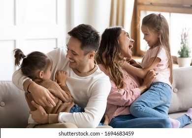 Smiling father cuddling small preschool daughter while happy mother tickling child girl, having fun together in modern living room. Laughing young family couple playing with little kids at home.