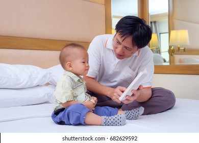 Smiling Father And Cute Little Asian 18 Months / 1 Year Old Toddler Baby Boy Reading Bedtime Story Book, Sitting On Bed At Home, Dad And Son Spending Time Together, Parents Reading To Child Concept