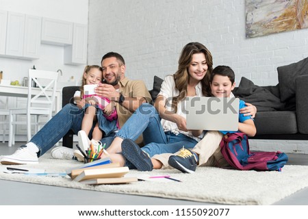 smiling father and daughter taking selfie on smartphone while mother and son using laptop together at home