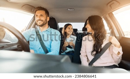 Smiling father focused on driving as his cheerful daughter in the backseat gives thumbs up, mother shares joyful moment looking towards her child