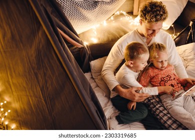 Smiling father and his cute kids wearing pajamas watching cartoons on digital tablet while sitting in handmade tent in children's room - Powered by Shutterstock