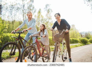 Smiling father and mother with daughter during summer outdoor bicycle riding. They enjoy togetherness in the summer city park. Happy parenthood and childhood or active sport life concept image. - Powered by Shutterstock