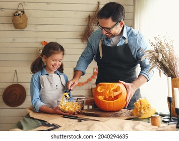 Smiling Father Showing His Little Daughter How To Carve Pumpkin With Spooky Face, Dad With Child Creating This Iconic Symbol Of Halloween Holiday Jack-o-lantern While Standing Behind Table In Kitchen