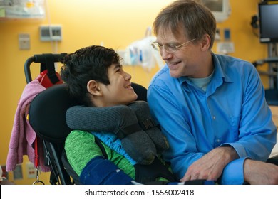 Smiling Father Sitting Next To Disabled Son In Wheelchair By  Hospital Bed,  Talking  Together