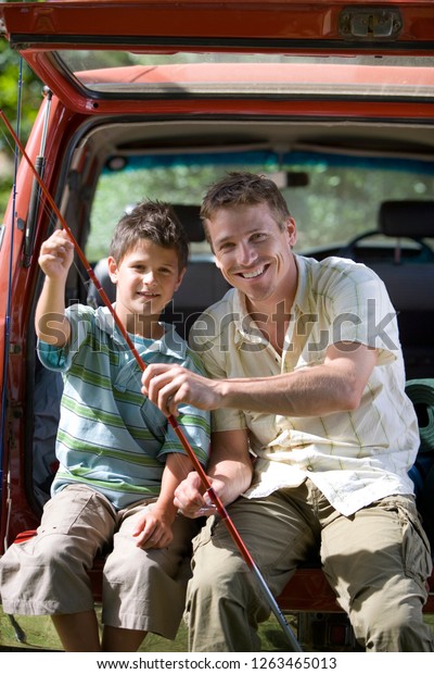 Smiling father and son in boot of car holding\
fishing rods at camera