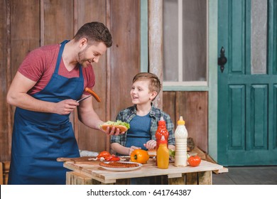 Smiling Father And Son Preparing Hot Dog Together In Backyard, Dad And Son Cooking Concept