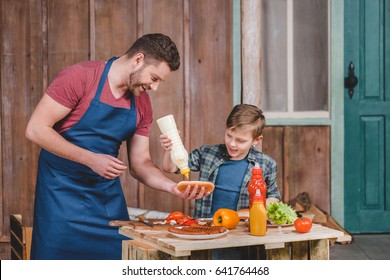 Smiling Father And Son Preparing Hot Dog Together In Backyard, Dad And Son Cooking Concept