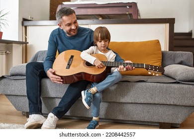 Smiling father teaching little son playing acoustic guitar on sofa at home. European family relationship and spending time together. Fatherhood and parenting. Domestic leisure, hobby and entertainment - Powered by Shutterstock