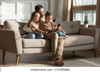 Smiling Father With Two Little Daughters Using Phone, Looking At Screen, Watching Cartoons Or Video, Playing Game, Shopping Online, Cute Preschool Girls With Dad Hugging, Sitting On Couch At Home