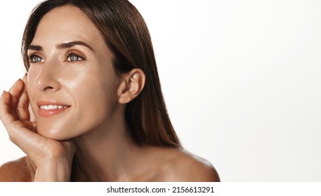 Smiling female aging model, clear glowing skin, nourished face without blemishes, applies daily cream, hyaluron anti-aging treatment, looking in mirror at herself, white background - Shutterstock ID 2156613291