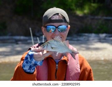 A Smiling Female Angler In Protective Sun Gear Holding Up A White Perch Fish In One Hand On A Sunny Day