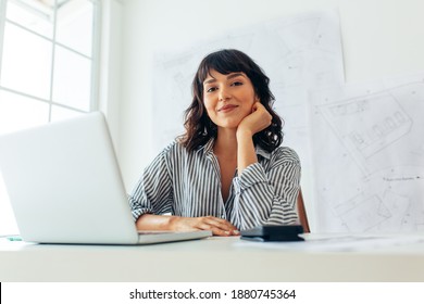Smiling female architect working on laptop in office. Entrepreneur sitting at workplace with architecture drawings in the background. - Shutterstock ID 1880745364