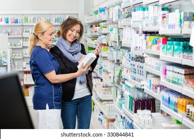 Smiling Female Assistant Showing Product To Customer Holding Cell Phone In Pharmacy