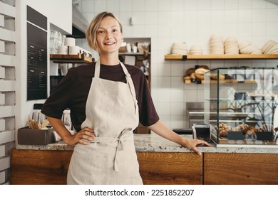 Smiling female baker entrepreneur standing at the counter of bakery and coffee shop. Young woman in cafe near showcase with fresh croissants and bread. Local small business owner indoors. - Powered by Shutterstock