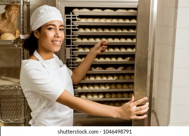 Smiling Female Baker Pointing At Dough Inside Of Industrial Oven At Baking Manufacture