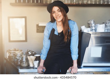 Smiling female barista sits on cafe counter wearing a black hat and apron over a long sleeved blue jean shirt besides rows of glass jars