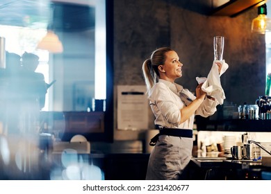 A smiling female bartender is wiping a glass while standing in a bar. - Powered by Shutterstock