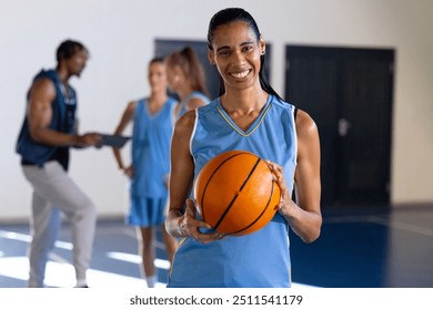 Smiling female basketball player holding ball in gym with teammates in background. Teamwork, sports, athlete, fitness, competition, camaraderie - Powered by Shutterstock