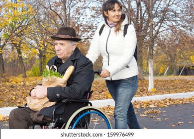 Smiling Female Carer Pushing An Old Handicapped Man In A Wheelchair Along The Street As They Return From Doing His Grocery Shopping