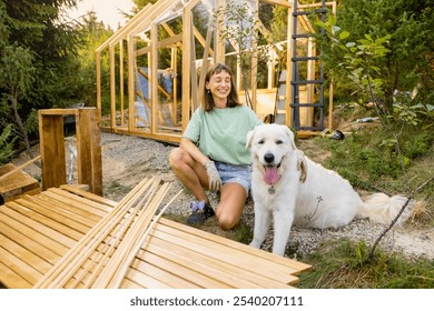 Smiling female carpenter kneeling next to her large white dog in front of a partially built wooden greenhouse in backyard, wearing casual clothes and work gloves. Concept of DIY project - Powered by Shutterstock