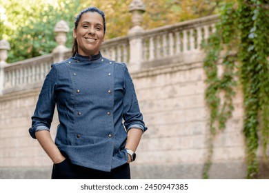 Smiling female chef wears blue coat, poses with hands on pockets outdoors on the street. Restaurant worker, culinary gourmet, pastry chef. - Powered by Shutterstock