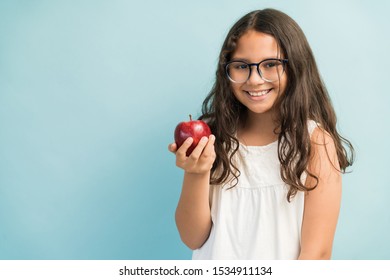 Smiling Female Child With Long Brown Hair Holding Fresh Apple While Making Eye Contact In Studio