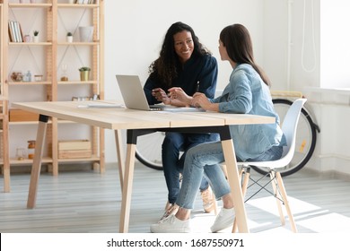 Smiling female colleagues sit at desk in office talk brainstorm work together on laptop, happy woman coworkers businesspeople speak discuss business ideas collaborate on modern computer at meeting