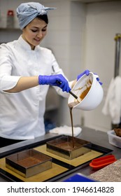 Smiling Female Confectioner Pouring Liquid Chocolate Gommage On Biscuit Cake In Metal Form Working At Catering Service Restaurant. Woman Chef In Uniform At Assembling Mousse Dessert Process