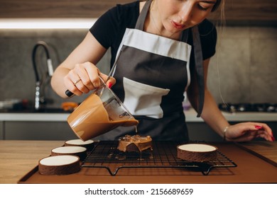 Smiling Female Confectioner Pouring Liquid Chocolate Gommage On Biscuit Cake. Woman Chef In Uniform At Assembling Mousse Dessert Process