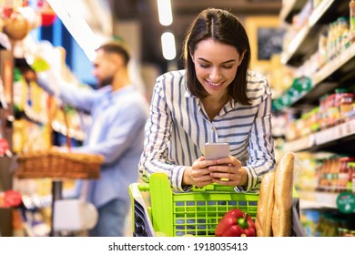 Smiling Female Customer Doing Grocery Shopping Using Smartphone Walking With Cart In Supermarket. Selective Focus. Woman Using Groceries Shopping Application On Phone Bying Food In Super Market