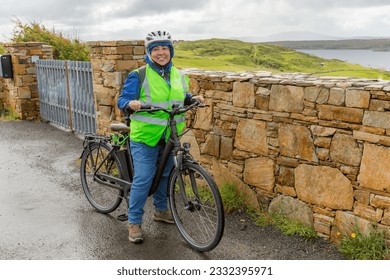 Smiling female cyclist pausing by fence, wearing jacket, helmet and reflective vest, Irish countryside and sea in background, cloudy rainy day in Clifden, County Galway, Ireland. Wild Atlantic Way - Powered by Shutterstock