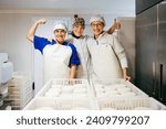 Smiling female dairy factory worker showing thumb up while embracing colleague demonstrating bicep standing near ready ricotta cheese after successful work