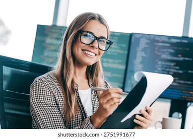 Smiling female developer holding a notepad and pen while reviewing code on multiple monitors in a bright modern office, conveying focus, confidence and collaborative software development.