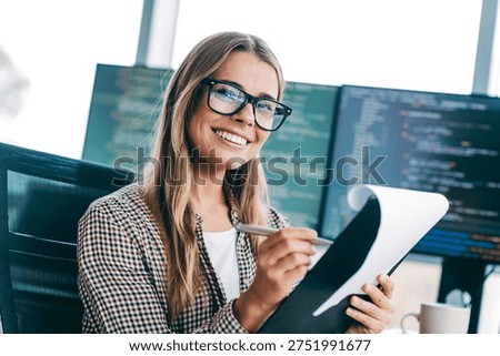 Smiling female developer holding a notepad and pen while reviewing code on multiple monitors in a bright modern office, conveying focus, confidence and collaborative software development.