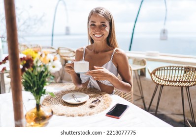 Smiling female digital nomad with long hair sitting at table with cup of coffee in hands and looking at camera in seaside cafe while travelling to Bali on summer holidays - Powered by Shutterstock