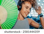 Smiling female dj wearing headphones is holding a green hand fan while playing music at a summer festival with bokeh lights in the background