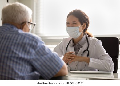 Smiling Female Doctor In Facemask And White Uniform Talk Consult Mature Male Patient At Consultation In Clinic. Woman GP In Facial Mask Have Meeting With Elderly Client. Healthcare, Covid-19 Concept.