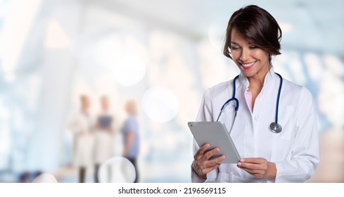 Smiling female doctor holding clipboard in her hand while standing at the clinic. Unrecognizable medical team standing at the background. Copy space. - Shutterstock ID 2196569115