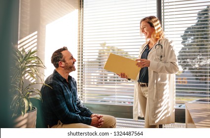 Smiling female doctor holding a medical report file and talking with male patient sitting on sofa at hospital reception. Doctor sharing good medical test results with the patient.