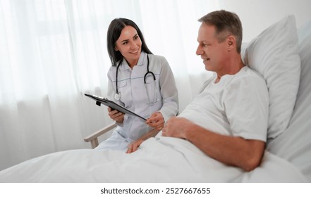 A smiling female doctor holds a clipboard while talking to a male patient in a hospital bed. The patient looks engaged and content. The bright, modern room emphasizes a caring medical interaction. - Powered by Shutterstock