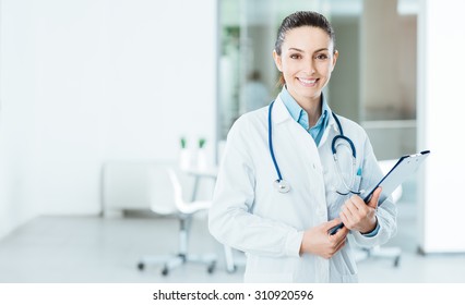 Smiling female doctor with lab coat in her office holding a clipboard with medical records, she is looking at camera