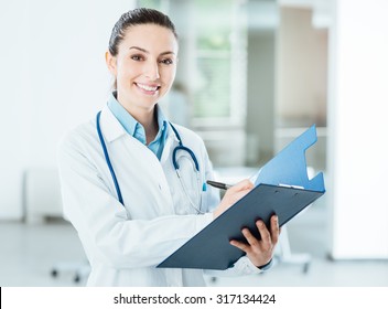 Smiling Female Doctor With Lab Coat In Her Office Holding A Clipboard With Medical Records, She Is Looking At Camera