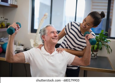 Smiling female doctor looking at senior male patient lifting dumbbells in hospital ward - Powered by Shutterstock