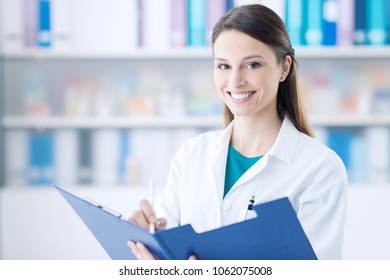 Smiling female doctor in the office holding a clipboard with medical records and writing